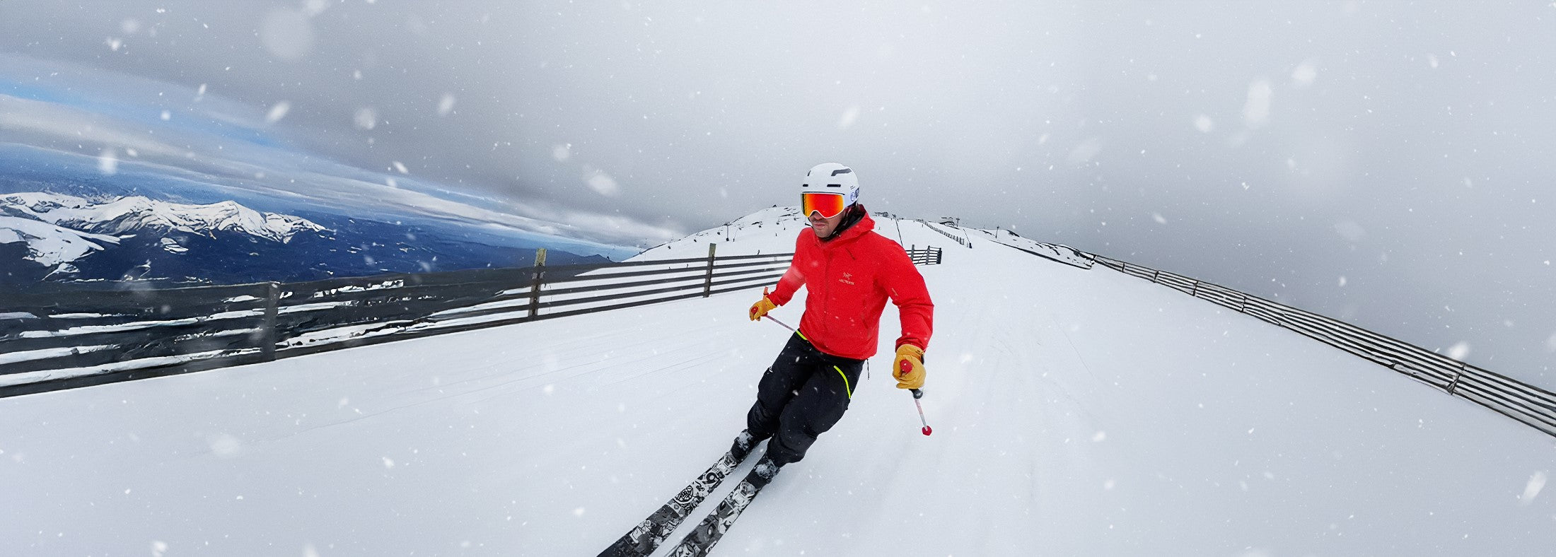 Skier in a red jacket carving down a snowy mountain slope during light snowfall with alpine scenery in the background.
