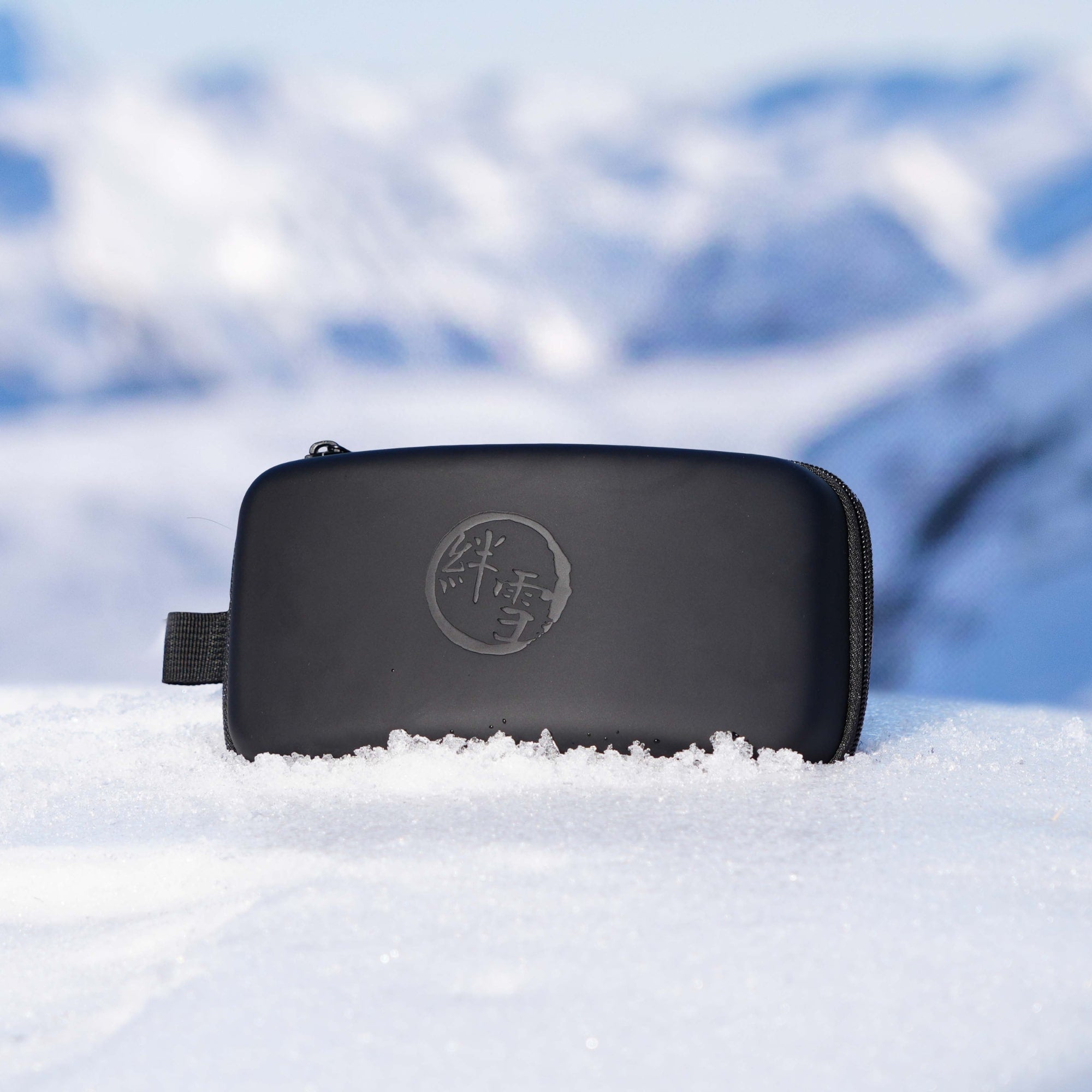 Black ski goggle case with a logo on it lying in the snow with a blurred mountain background