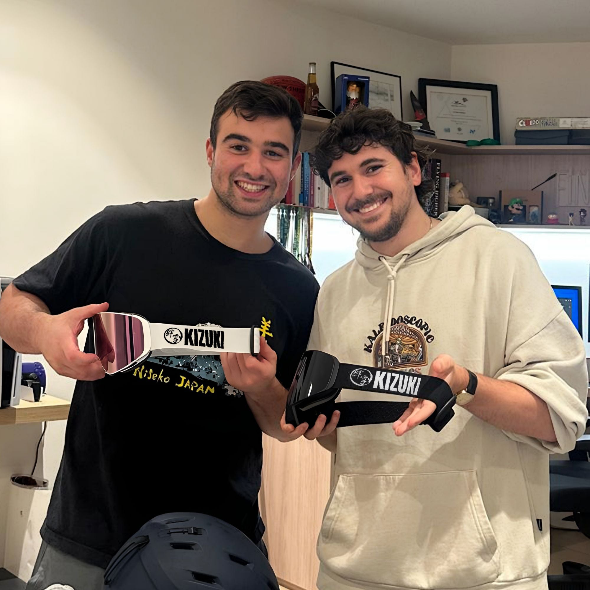 Two men smiling indoors while holding KIZUKI ski goggles, showcasing different lens styles and strap designs.