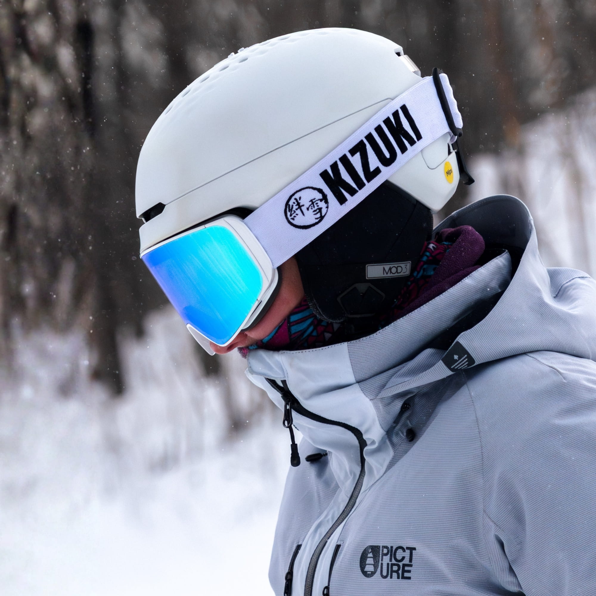 Female wearing a blue ski goggle with white strap, in a snowy setting.