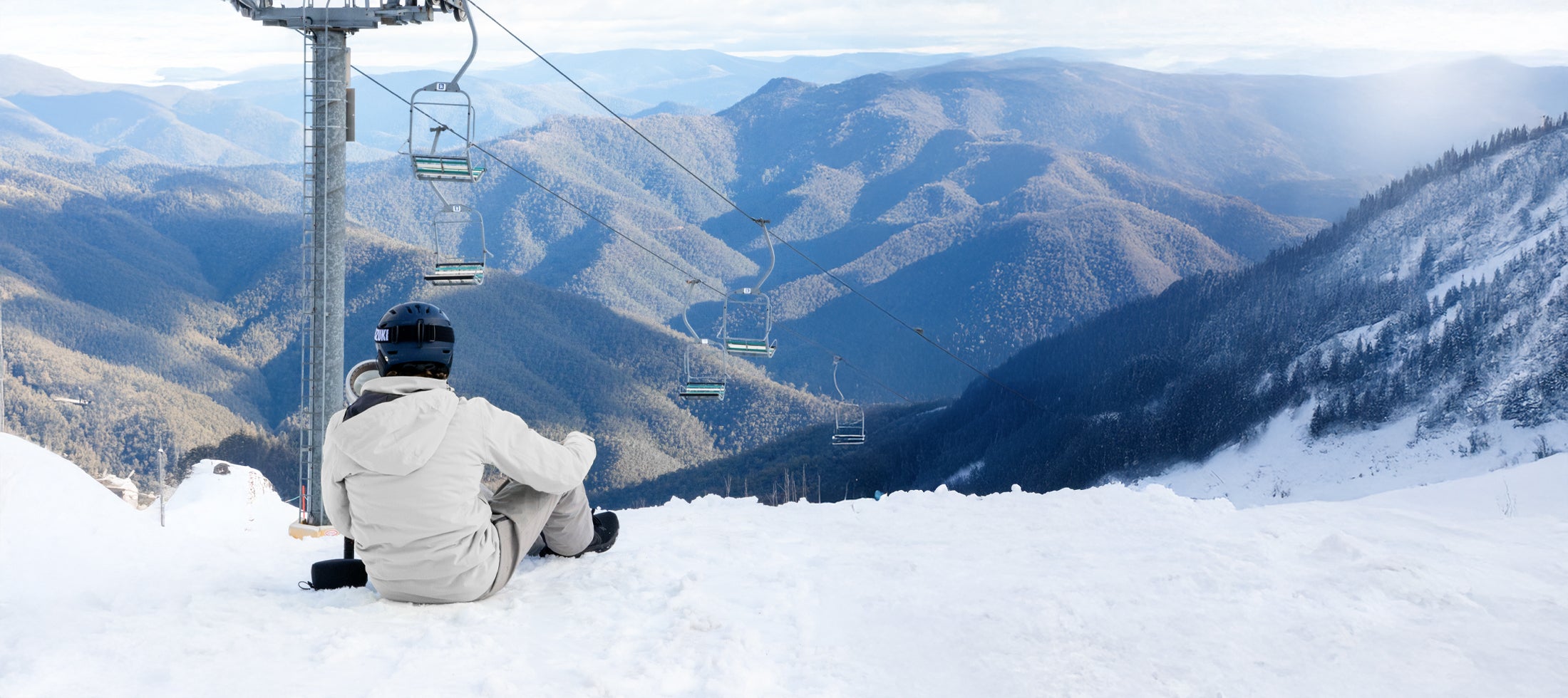 Skier sitting on a snowy slope overlooking alpine mountains with a chairlift running across the landscape.