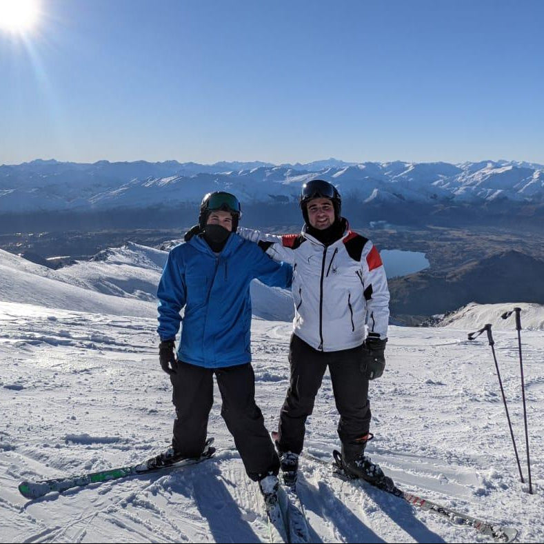 Two skiers standing together on a snowy mountain slope under a bright sun, surrounded by panoramic alpine peaks and a clear blue sky.