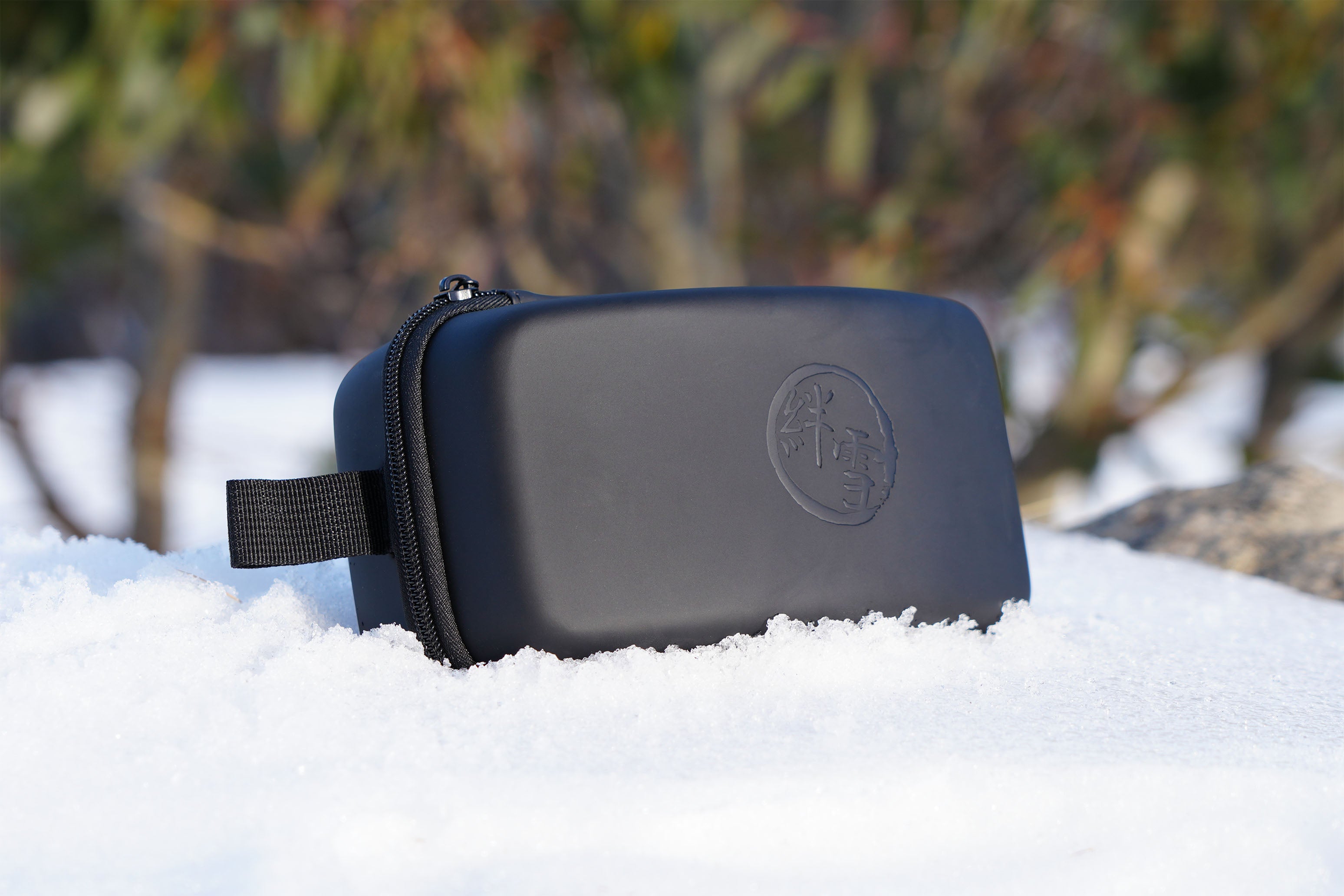 Black ski goggle case sitting in the snow with green bushes behind