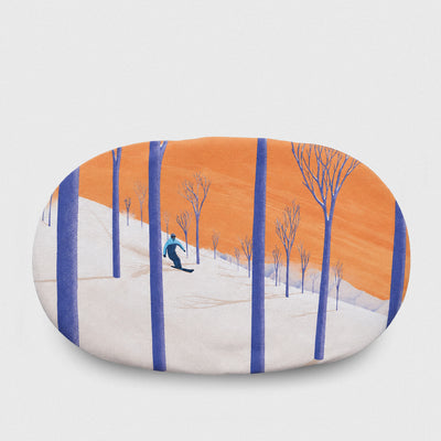 An oval-shaped goggle cover featuring a stylized winter landscape illustration with a skier on a snowy slope, blue tree trunks, and a warm orange sky, shown against a light gray background.