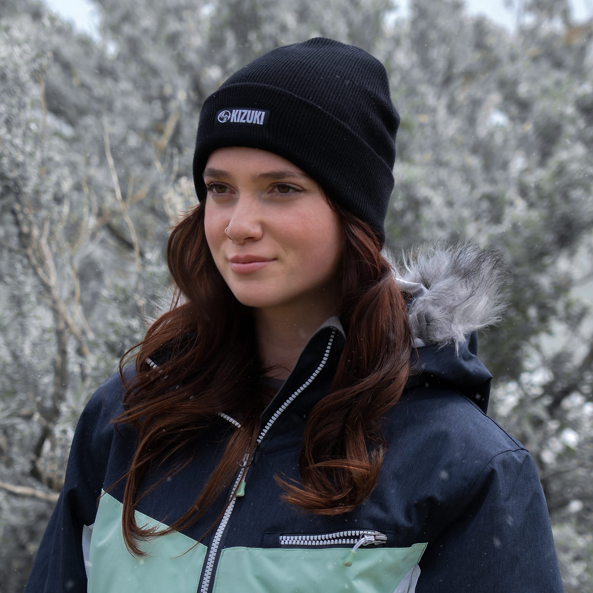 Person wearing a black knit beanie with a brand logo, standing outdoors against a snowy background.