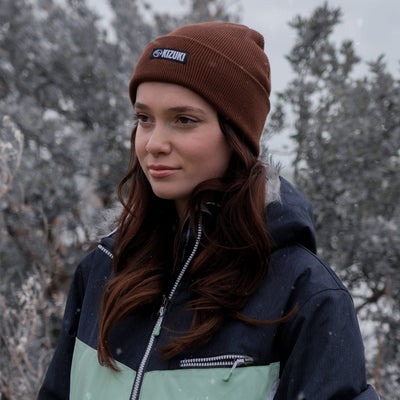 Person wearing a brown knit hat with a logo and a dark jacket in a snowy forest.