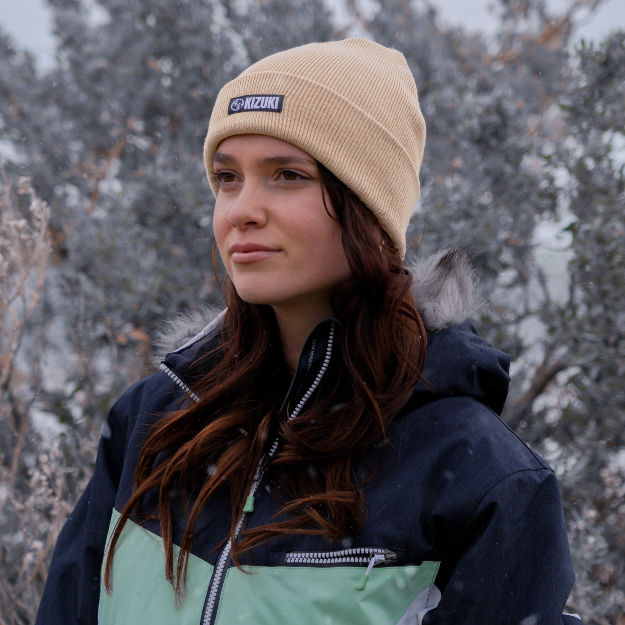 Woman wearing a beige beanie with a logo and a dark jacket in a snowy landscape.