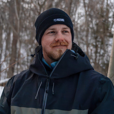 Man wearing a black jacket and beanie with a brand logo, standing in a forested area.