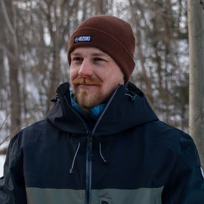 Man wearing a brown knit cap and dark jacket standing in a forested area.