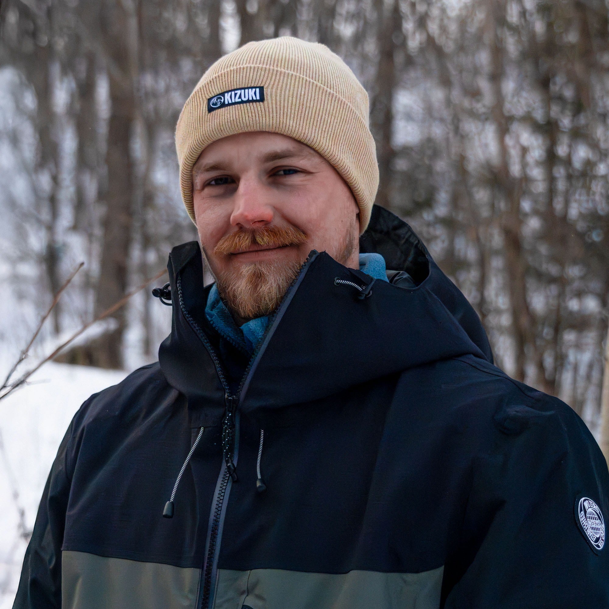 Man wearing a beige knit cap with a brand logo and a dark jacket in a snowy forest.