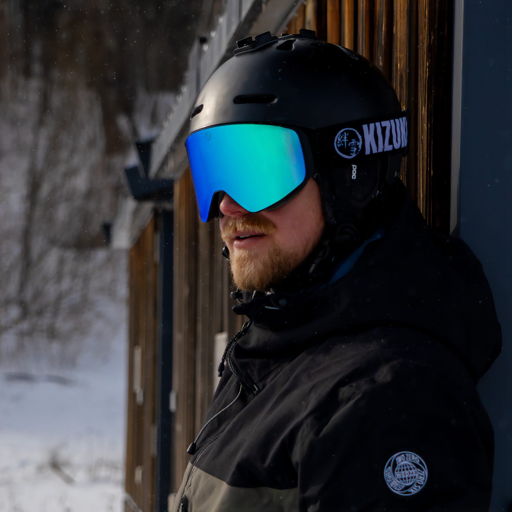 Man standing near a cabin wearing KIZUKI Dawn ski goggles with Glacial Blue lens and black helmet.