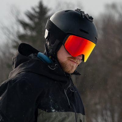 Man wearing KIZUKI Dawn ski goggles with Horizon Red lens and black helmet in winter landscape.