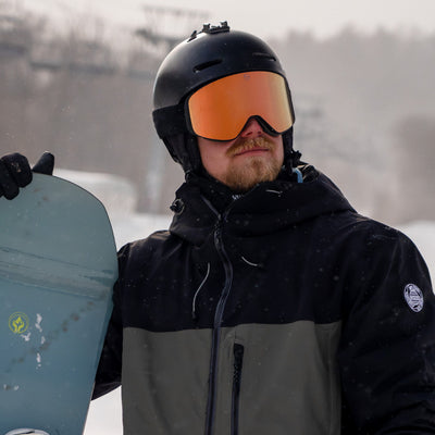 Man holding a snowboard wearing KIZUKI Dawn ski goggles with Twilight Rose lens and black helmet.