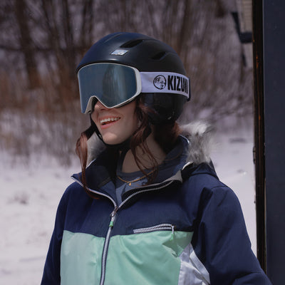 Woman smiling while wearing KIZUKI Dawn white frame ski goggles with Blizzard Silver lens and black helmet in snowy setting.