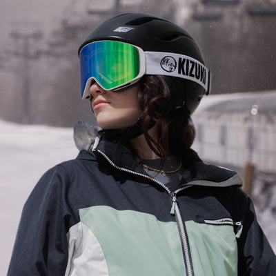Woman wearing KIZUKI Dawn white frame ski goggles with Forest Green lens and black helmet on snowy slopes.
