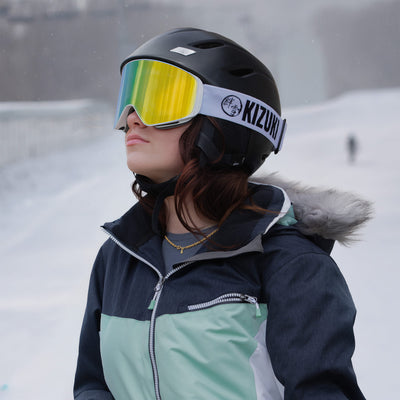 Woman wearing KIZUKI Dawn white frame ski goggles with Summit Gold lens and black helmet in alpine setting.
