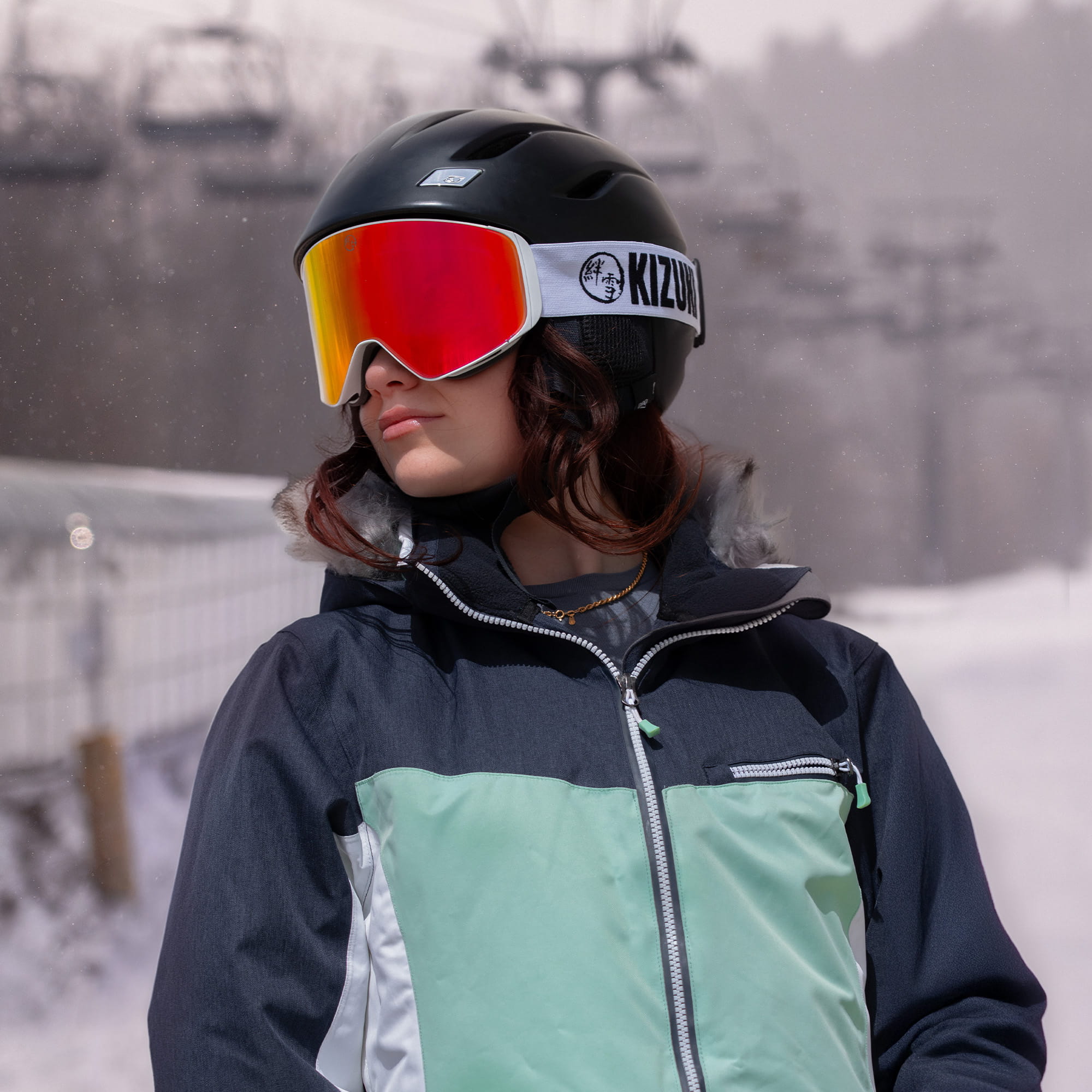 Woman wearing KIZUKI Dawn white frame ski goggles with Horizon Red lens and black helmet outdoors in snow.