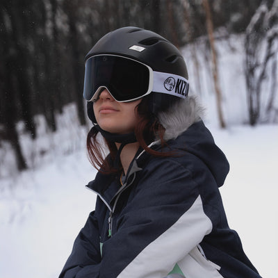 Woman wearing KIZUKI Dawn white frame ski goggles with Midnight Black lens and black helmet in winter landscape.