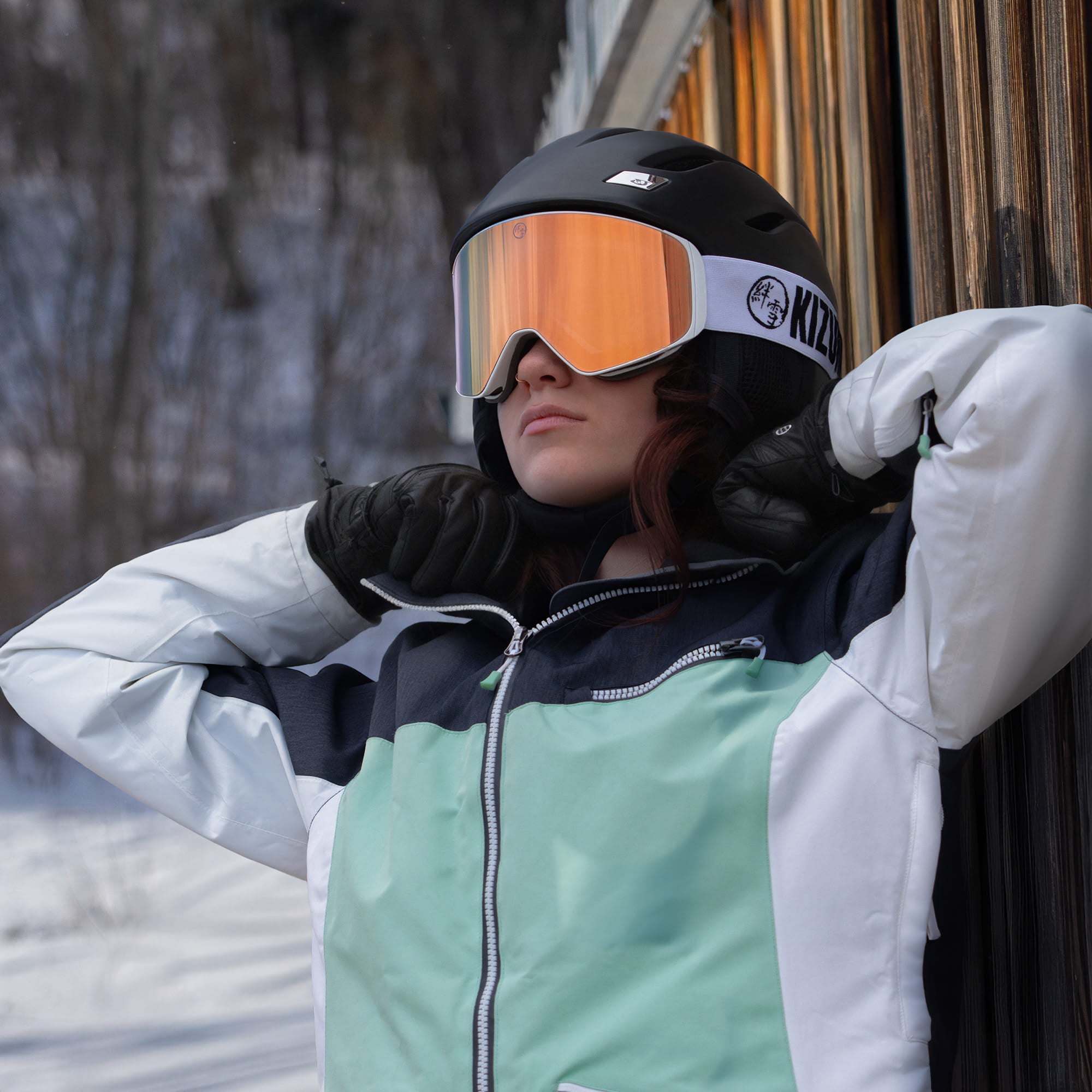 Woman leaning against a wooden wall wearing KIZUKI Dawn white frame ski goggles with Twilight Rose lens and black helmet.