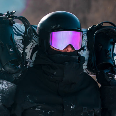 Person wearing a black helmet with pink goggles and dark clothing, sitting on a snowboard with a blurred background.