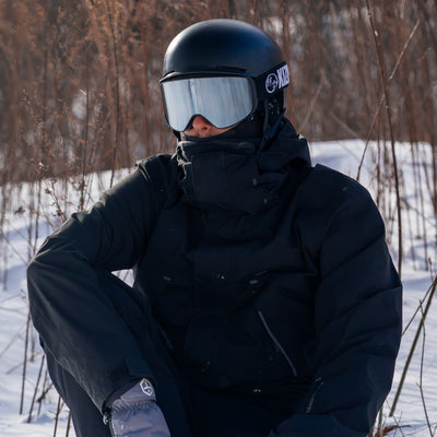 Person in black winter gear & silver goggles, sitting in a snowy landscape