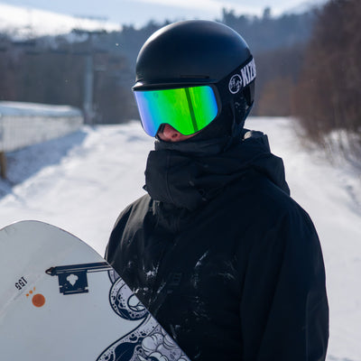 Person wearing a black helmet with green goggles and holding a snowboard in a snowy landscape.