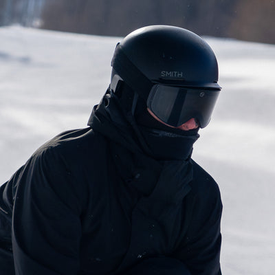 Person wearing a black helmet and jacket with black ski goggles in a snowy landscape