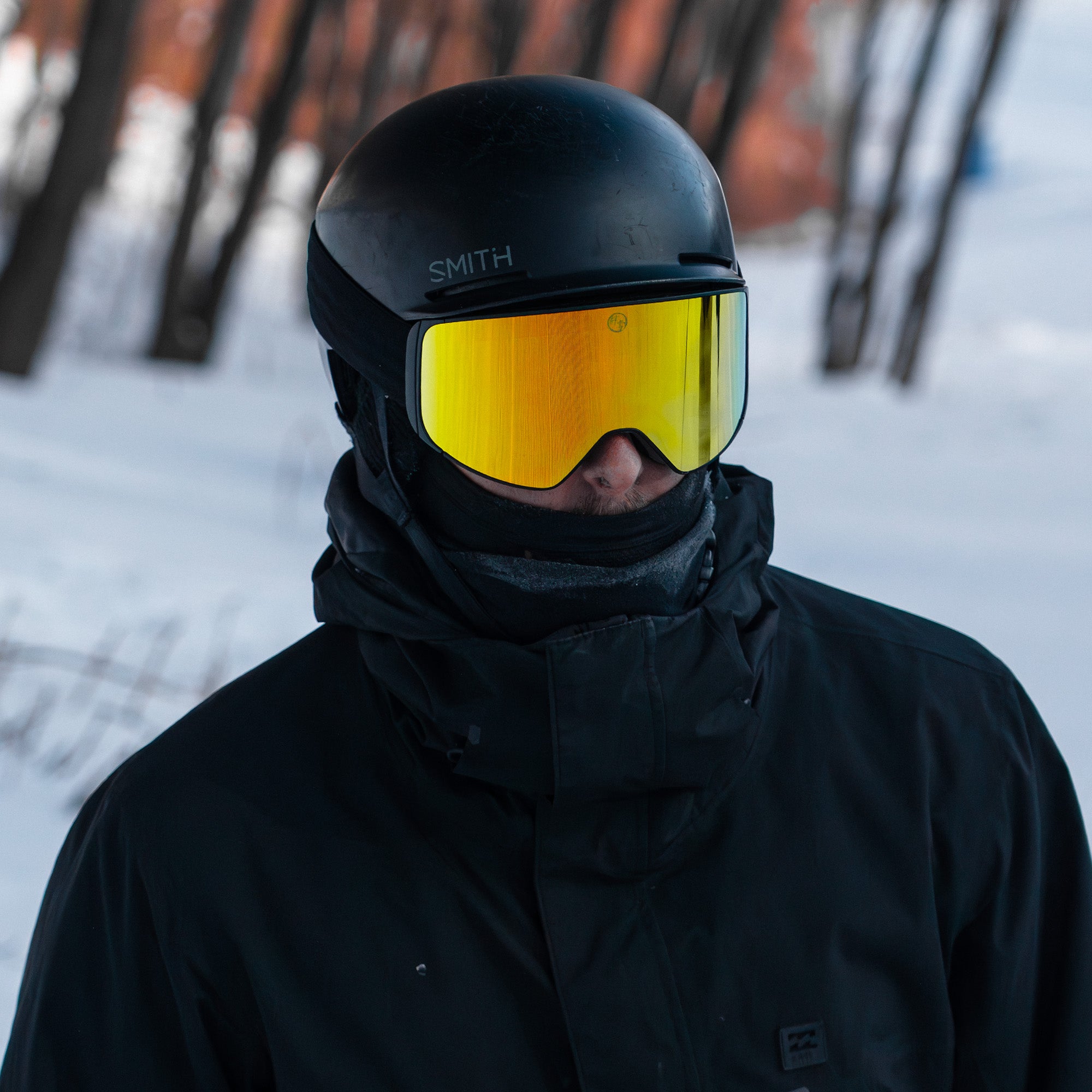 Person wearing a black helmet with gold goggles and black jacket in a snowy setting