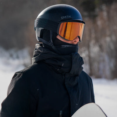 Person wearing a black helmet with rose gold goggles and black jacket in a snowy landscape