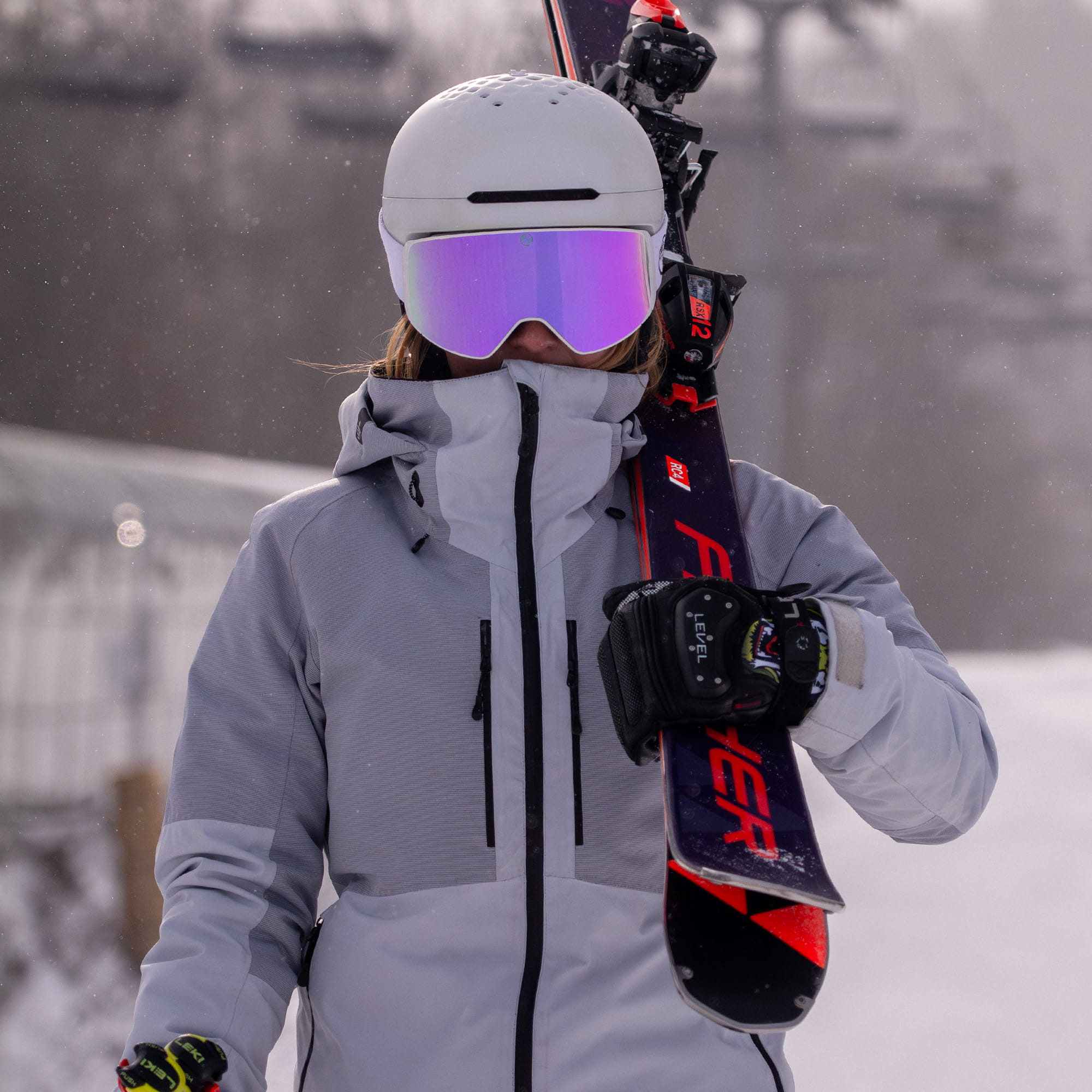 Person in ski gear, wearing pink goggles holding skis with a blurred background