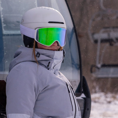 Person in winter gear with helmet and green goggles standing in front of a vehicle.