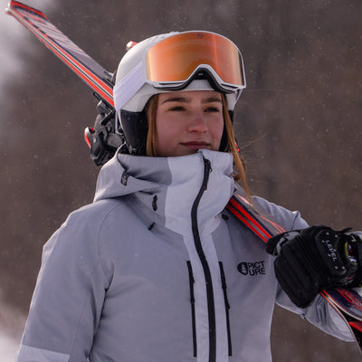 Person wearing a gray jacket with a brand logo, holding skis and rose gold goggles in a snowy environment.