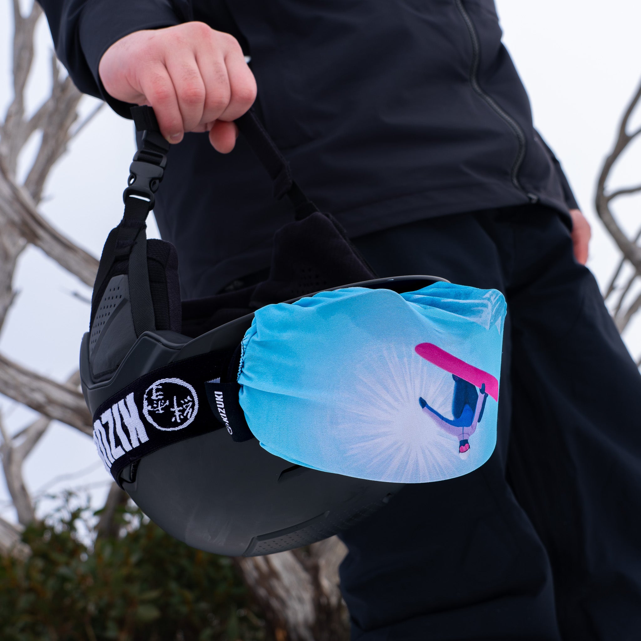 A person outdoors wearing a black ski jacket and pants, carrying a black ski helmet with a bright blue and pink printed goggle cover attached to a strap.