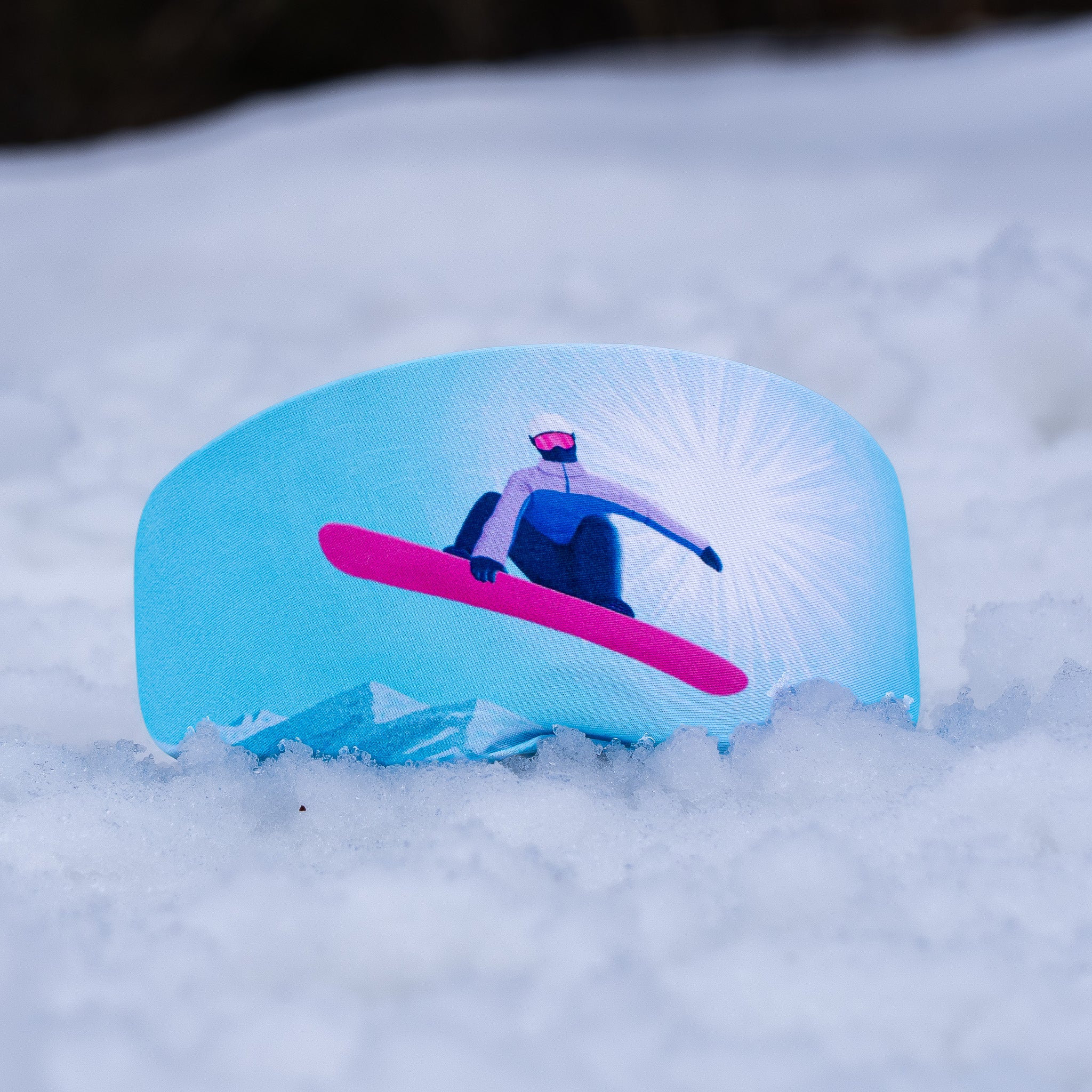 A light blue curved product, goggle cover, printed with an illustration of a snowboarder on a bright pink board, set in deep snow.
