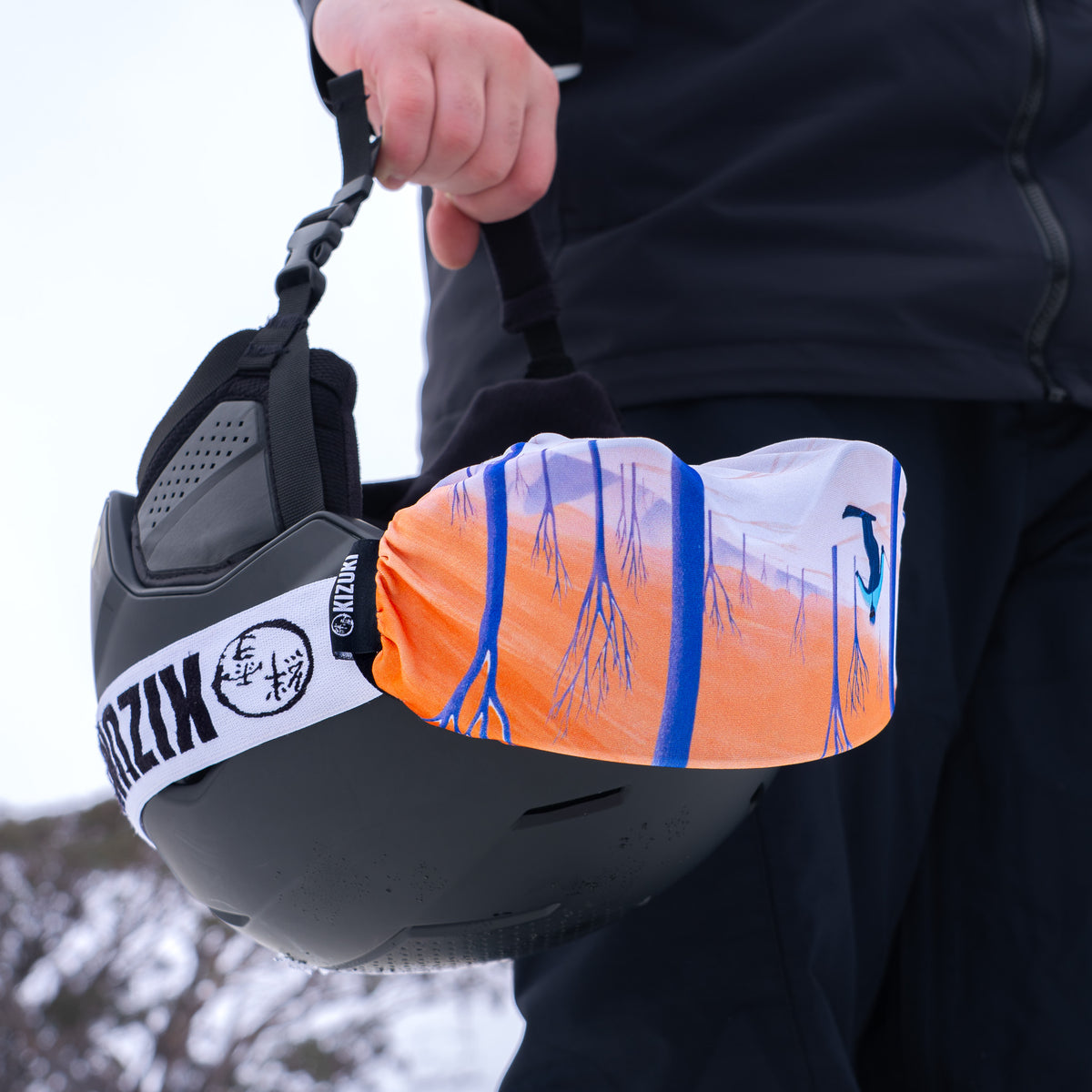 A close-up of a person outdoors holding a black ski helmet with a bright orange, white, and blue printed snowboard sleeve or cover draped over the front.