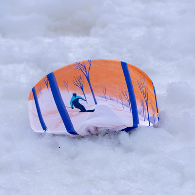 A goggle cover with a printed winter landscape design, featuring an orange sky, blue tree trunks, and a skier, resting on a bed of white snow.