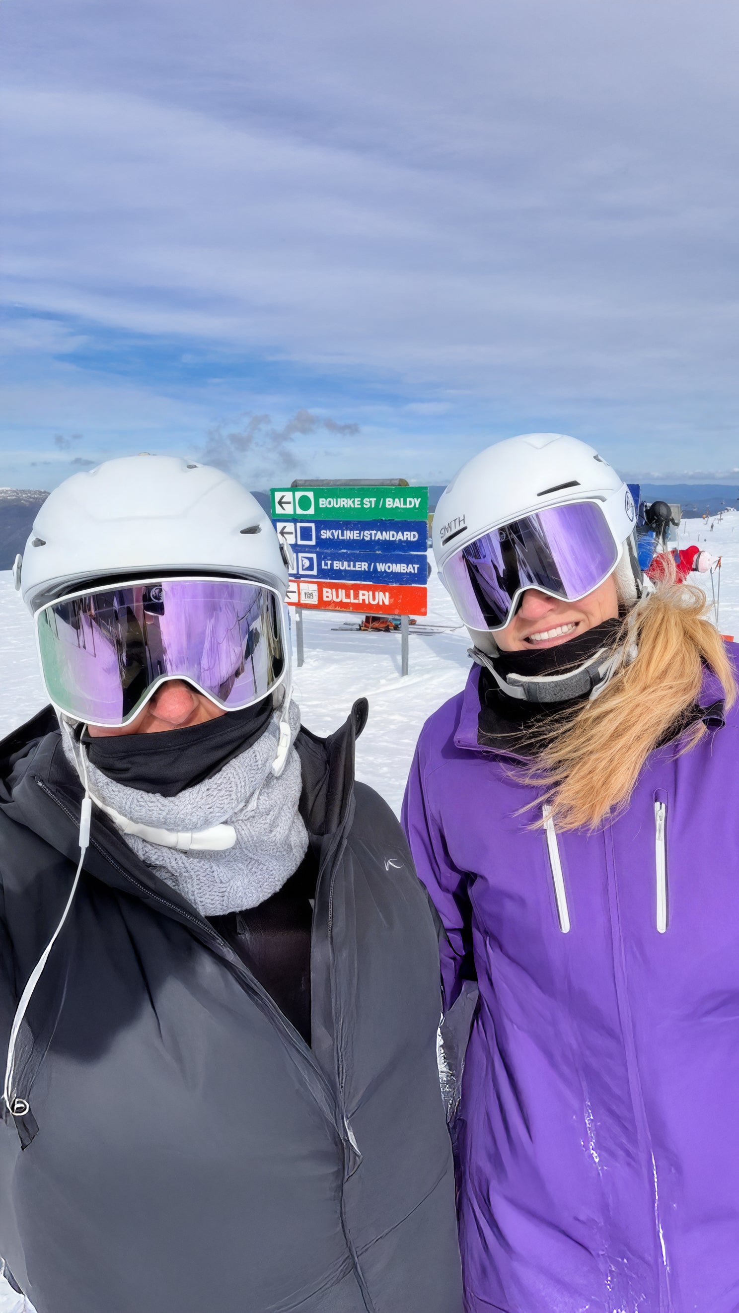Two people in ski gear standing on a snowy slope with ski resort signs in the background.